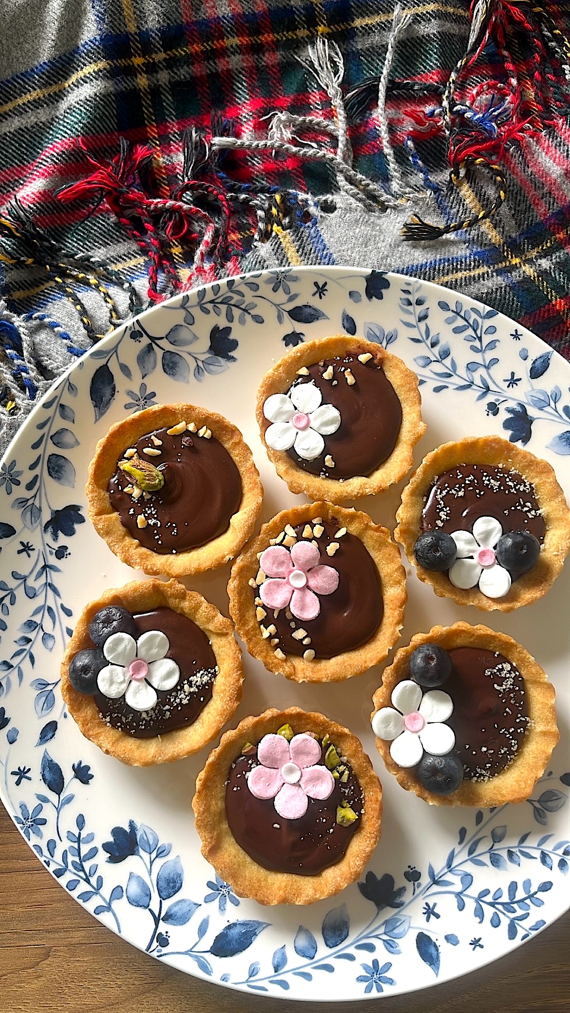 Seven small chocolate tarts decorated with flower-shaped sugar toppings and blueberries